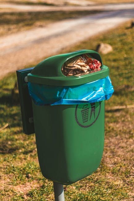A small outdoor public rubbish bin located on a grassy area, featuring a rounded green plastic body with a semi-transparent, curved lid made of dark green material. The lid is slightly open, revealing